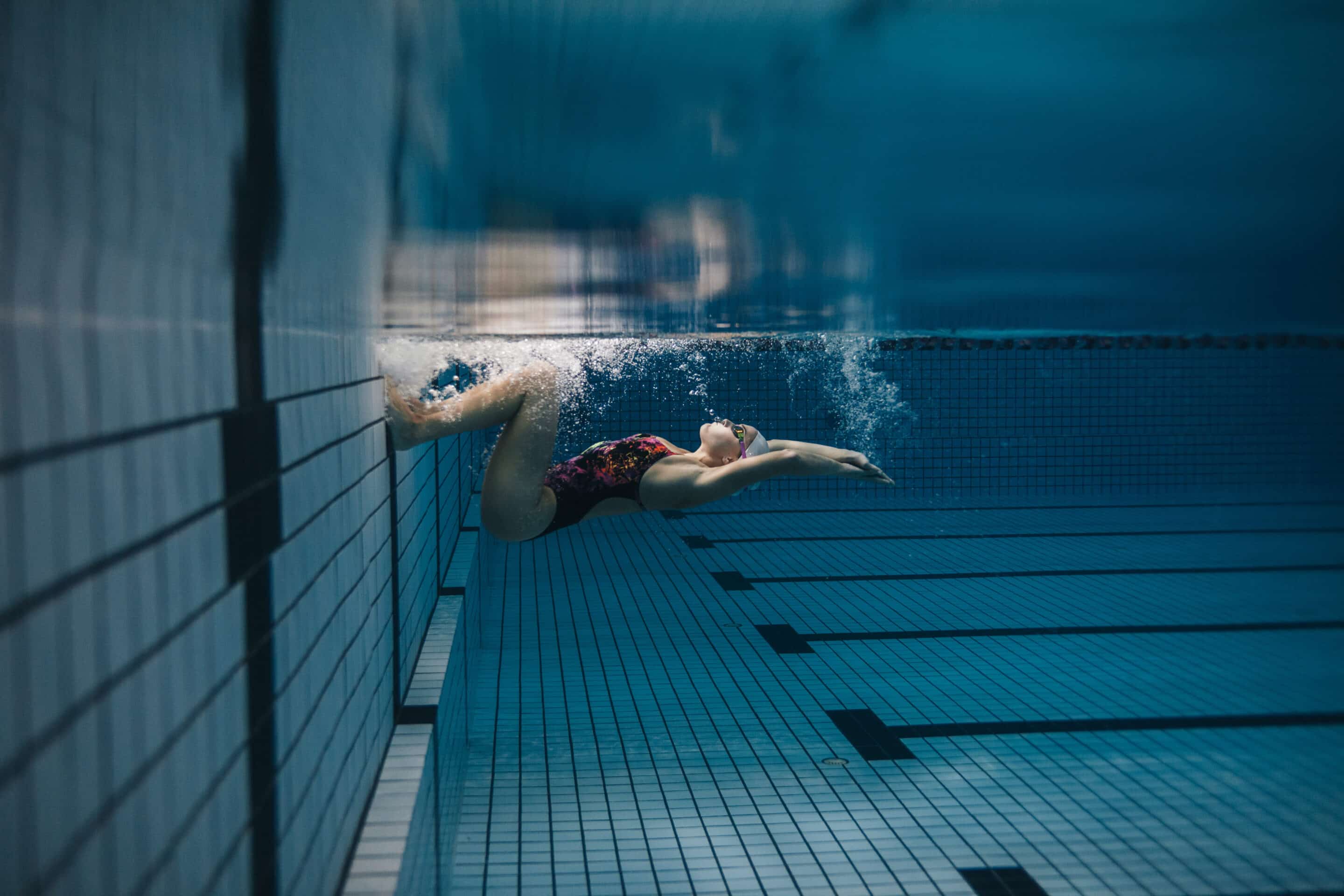 Underwater image of a swimmer gliding along the blue tiled pool floor beside the wall.